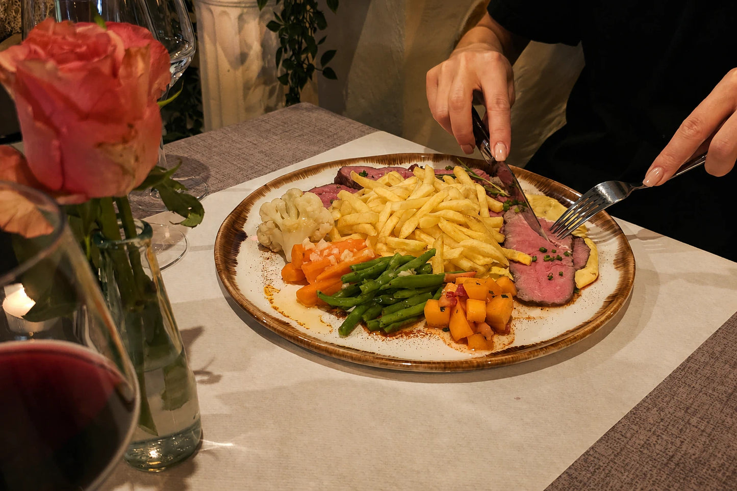 Person schneidet zartes Roastbeef mit Pommes frites und Gemüse auf einem stilvoll angerichteten Teller im Restaurant an.